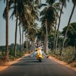 Un couple joyeux conduit un scooter jaune sur une route pittoresque bordée de palmiers, capturant l'essence du voyage et de l'aventure tropicale.